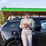 Une jeune femme blonde avec un smartphone dans une station de recharge pour charger une voiture électrique à proximité à Chisinau, en Moldavie