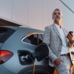Un homme d'affaires détenant un smartphone tout en chargeant une voiture à la station de charge de véhicules électriques, Clatep.